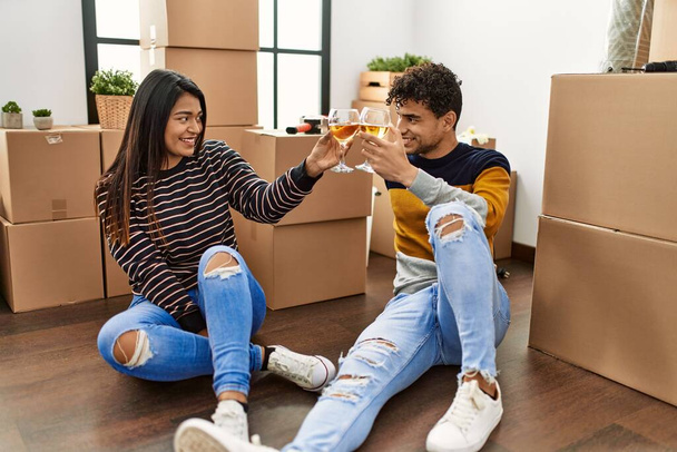 Junges lateinisches Paar stößt im neuen Zuhause mit Wein auf dem Fußboden an. - Foto, Bild