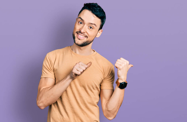 Homme hispanique à la barbe portant un t-shirt décontracté pointant vers le dos derrière avec la main et les pouces levés, souriant confiant  - Photo, image