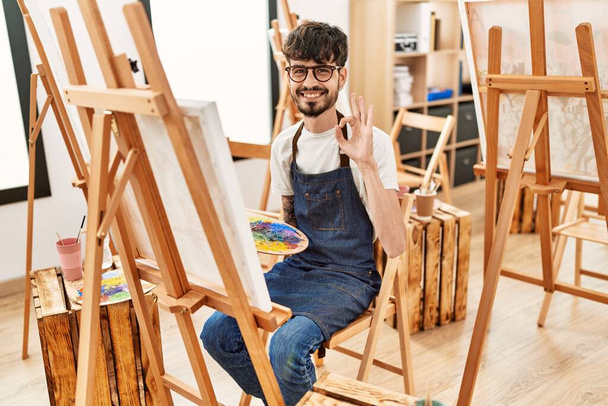 Hispanischer Mann mit Bart im Kunststudio macht ok Zeichen mit den Fingern, lächelt freundlich gestikulierend hervorragendes Symbol  - Foto, Bild