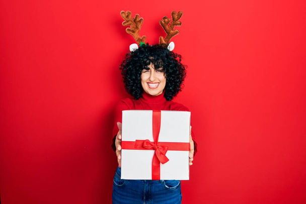 Young middle east woman wearing deer christmas hat holding gift winking looking at the camera with sexy expression, cheerful and happy face.  - Photo, Image