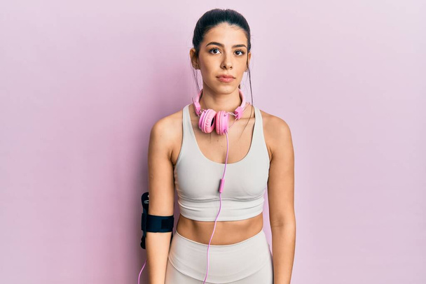 Young hispanic woman wearing gym clothes and using headphones relaxed with serious expression on face. simple and natural looking at the camera.  - Photo, Image
