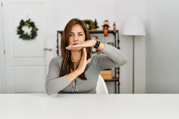 Mujer latina joven sentada en la mesa por la decoración de Navidad haciendo tiempo fuera gesto con las manos, rostro frustrado y serio  - Foto, Imagen