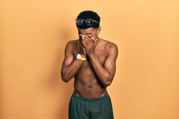 Junger afrikanisch-amerikanischer Mann in Badebekleidung und Schwimmbrille mit traurigem Gesichtsausdruck, der weinend das Gesicht mit den Händen bedeckt. Depressionskonzept.  - Foto, Bild