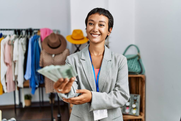 若いヒスパニック系女性の店主笑顔自信を持って保持ドル紙幣で衣料品店 - 写真・画像