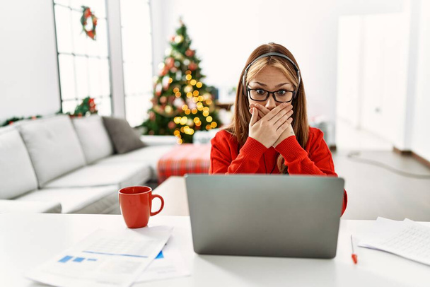 Mladá běloška sedící na stole a používající notebook u vánočního stromku, šokovaná, zakrývající ústa rukama, aby se nespletla. tajný koncept.  - Fotografie, Obrázek