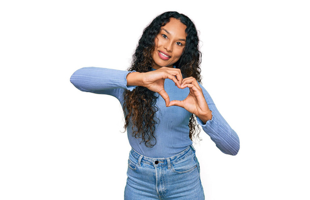 Jonge Latijns-Amerikaanse vrouw met krullend haar in casual kleding glimlachend in liefde doen hart symbool vorm met handen. romantisch concept.  - Foto, afbeelding