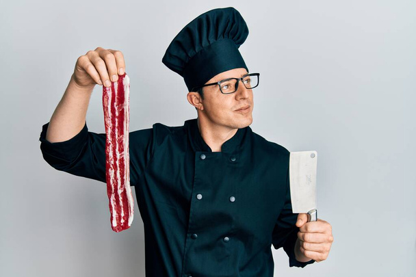 Guapo joven con uniforme de chef sosteniendo tocino y cuchillo sonriendo mirando a un lado y mirando hacia otro pensando.  - Foto, Imagen