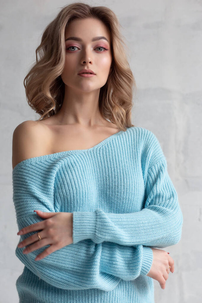 Sexy blonde girl in a blue knitted sweater on a white wall background. Fashion photo of a gorgeous close-up model - Фото, зображення