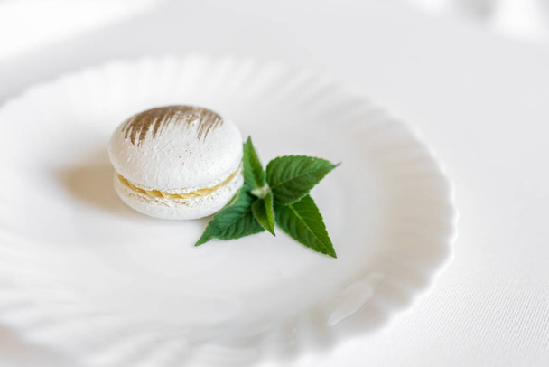 White macarons with a green mint leaf on a white plate on a light background. Delicious and beautiful pastries. Background for confectionery - Fotoğraf, Görsel