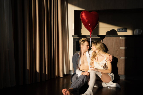 ευτυχισμένο ζευγάρι αγάπη άντρας και γυναίκα πρωί στις ακτίνες του ήλιου κάθονται στην κουζίνα κρατώντας ένα μπαλόνι σε σχήμα καρδιάς στα χέρια τους. έννοια της γιορτής ημέρα του Αγίου Βαλεντίνου, επέτειο ή επέτειο. - Φωτογραφία, εικόνα