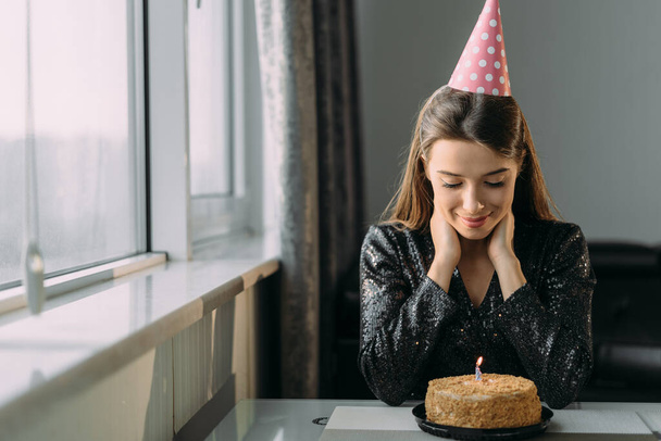Portrait of a sad young woman sitting at the table on her birthday, a lonely holiday at home, a girl in a cap celebrating an anniversary in quarantine, the concept of celebrating a birthday - Photo, Image