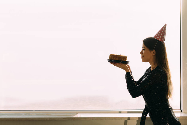 beautiful caucasian woman holding a cake looking at a candle, standing at the office window, light background, profile view of a girl with a birthday cake, birthday celebration concept, space for text - 写真・画像