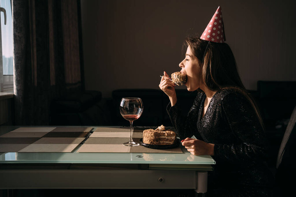 Una mujer comiendo un pastel abriendo la boca, una niña de 25 años celebra su cumpleaños, una morena sentada en la mesa con una gorra festiva. concepto de casa de vacaciones - Foto, Imagen