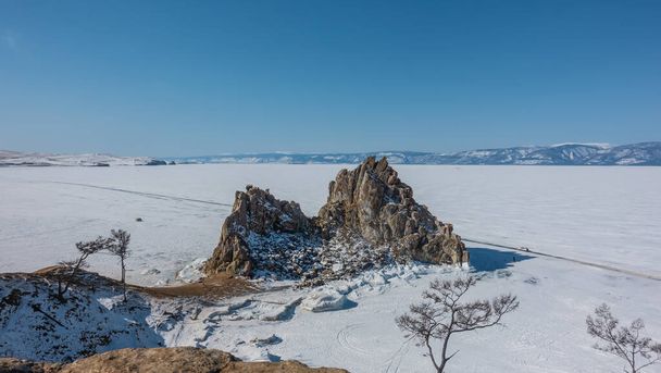 Malebná dvouhlavá skála na pozadí zamrzlého jezera. Prudké svahy jsou pokryty sněhem. Auto jede po ledu. Horské pásmo proti modré obloze. Holé stromy. Bajkal. Shamanka  - Fotografie, Obrázek