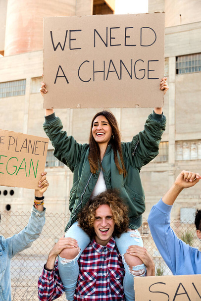 Feliz joven hispana sentada sobre los hombros de los hombres protestando contra el cambio climático en manifestación. Imagen vertical. - Foto, imagen