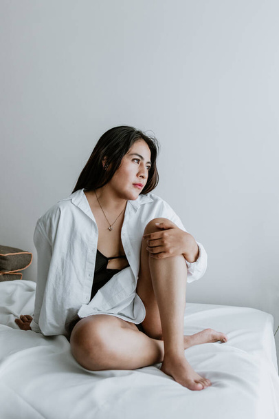 Young Latin woman sitting on bed, wearing white robe, showing legs at home in Mexico city - Фото, изображение