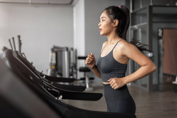 Asian woman exercises in fitness. Young healthy woman in sportswear is running on a treadmill in gym. Exercises and fitness concept. - Photo, Image