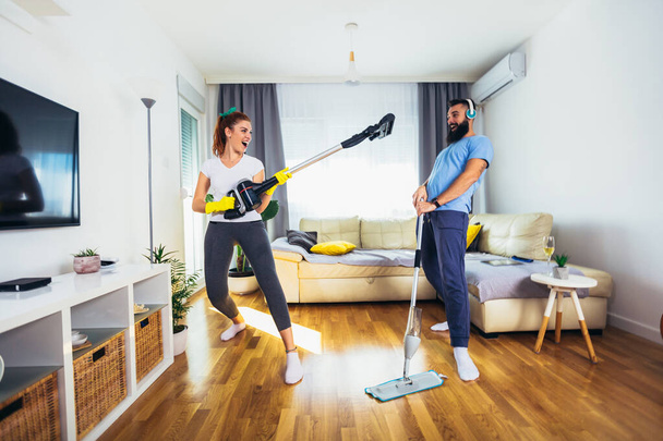 Smiling excited young housewife and her husband having fun with vacuum cleaner - Foto, Imagen