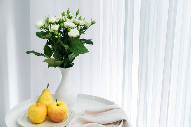 Vase avec bouquet de belles roses fraîches et table de fruits dans la chambre - Photo, image
