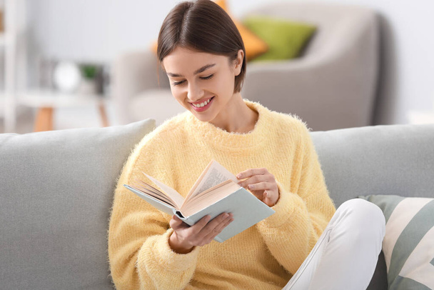 Beautiful young woman reading book on sofa at home - Foto, Imagem