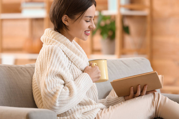 Beautiful young woman drinking tasty tea and reading book on sofa at home - Photo, Image