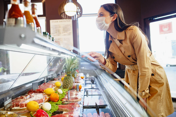 Szczęśliwa młoda kobieta w masce do twarzy wybierając mięso ze szklanej szafki w sklepie spożywczym - Zdjęcie, obraz
