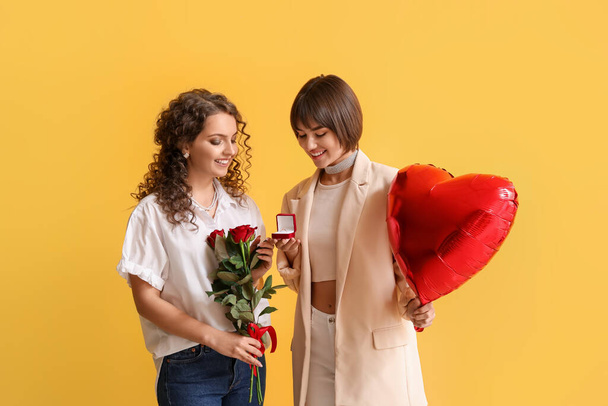 Happy lesbian couple with engagement ring, flowers and balloon on color background - Photo, Image