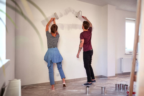 Joven pareja caucásica pintando la forma de la casa en la pared en casa nueva - Foto, Imagen