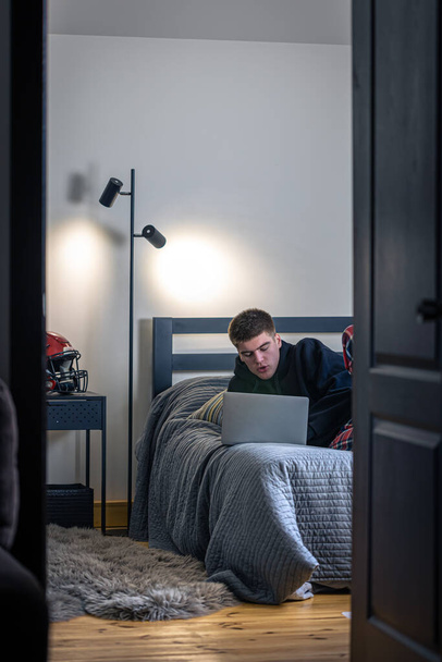 A teenager guy sits in a room on a bed and uses a laptop. - Photo, Image