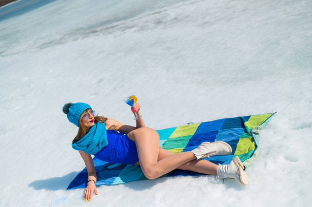 Beautiful caucasian woman in a swimsuit and a hat sunbathes and drinks a cocktail on the snow.  - Foto, Imagem