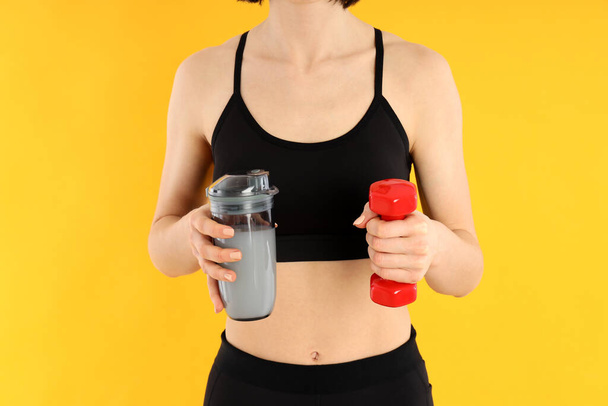 Αθλητική γυναίκα κατέχει πρωτεΐνη κοκτέιλ και αλτήρα σε κίτρινο φόντο - Φωτογραφία, εικόνα