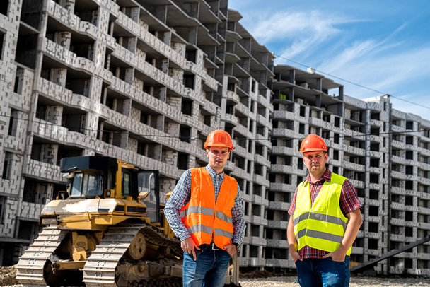 two strong young workers in uniform stand on the background of a new building. The concept of a new building - Zdjęcie, obraz
