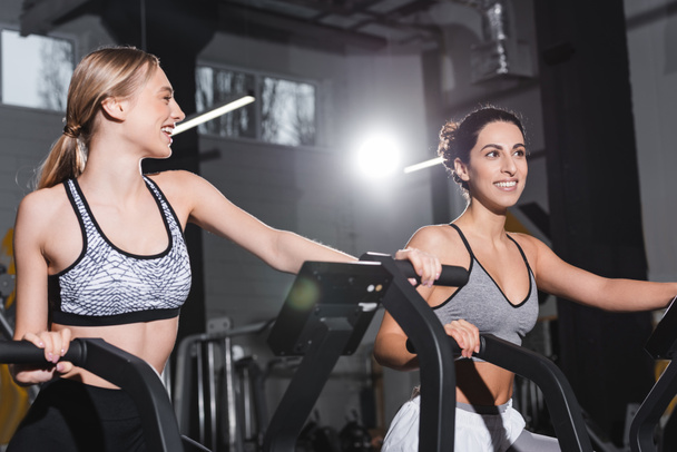 Cheerful interracial women training on elliptical trainers in sports center  - Foto, Imagem