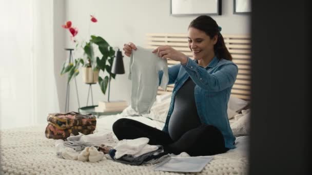 Biała kobieta w zaawansowanej ciąży, siedząca na łóżku i składająca ubrania dla dziecka do szpitala. Nakręcony aparatem hel RED w rozdzielczości 8K. - Materiał filmowy, wideo