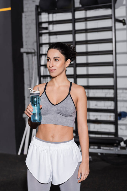 Smiling middle east sportswoman holding sports bottle in gym  - Foto, imagen