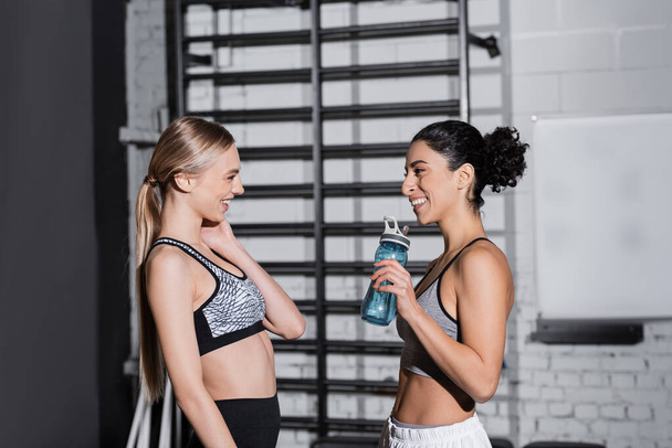 Side view of positive interracial sportswomen with sports bottle talking in gym  - Valokuva, kuva