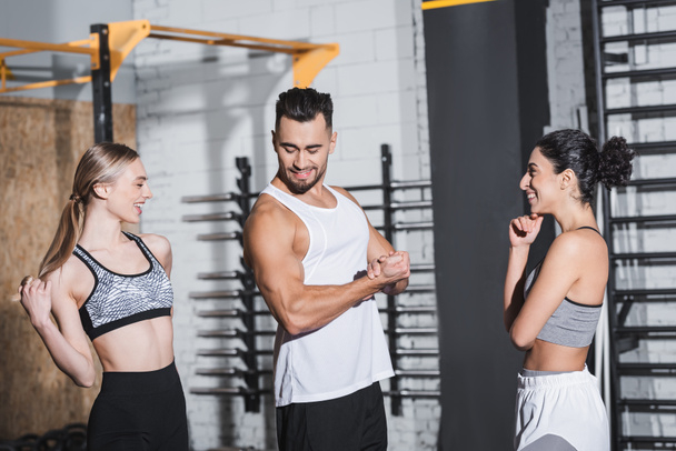 Smiling sportsman showing muscles near interracial sportswomen in gym  - 写真・画像