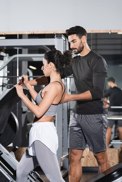 Arabian trainer standing near middle east sportswoman and leg press machine in gym  - Foto, afbeelding
