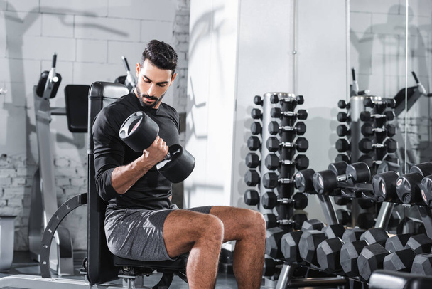 Muslim sportsman looking at dumbbell while training in gym  - Fotó, kép