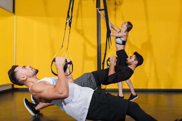 Vue latérale du sportif tirant des bretelles de suspension près d'amis interraciaux sur fond flou dans la salle de gym  - Photo, image