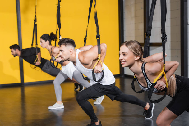 Blonde sportswoman training with suspension straps near blurred interracial friend in gym  - Fotó, kép