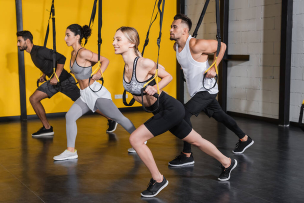 Sportive positive tirant des bretelles de suspension près de personnes multiethniques dans la salle de gym  - Photo, image