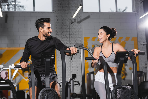 Smiling arabian sportsman looking at middle east sportswoman on elliptical trainer in gym  - Foto, Bild