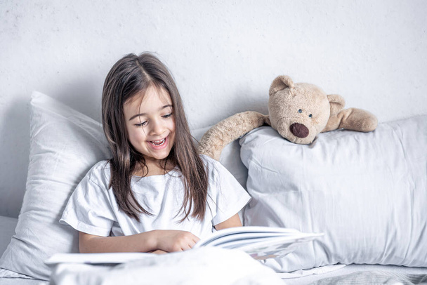 Маленька дівчинка читає книгу з плюшевим ведмедем в ліжку вранці
. - Фото, зображення