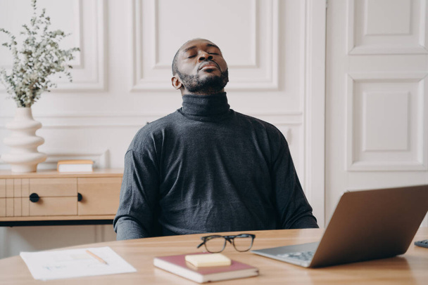 Εξαντλημένος υπάλληλος γραφείου Αφρο-Αμερικανός άνδρας κάθεται μπροστά από το laptop βγάζοντας γυαλιά κλείνοντας τα μάτια και πετώντας το κεφάλι πίσω. Κουρασμένος νεαρός άνδρας προσπαθεί να ανακτήσει για να συνεχίσει την απομακρυσμένη εργασία σε απευθείας σύνδεση - Φωτογραφία, εικόνα