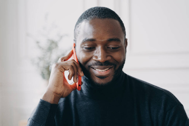Primer plano de un joven africano sonriente que tiene una conversación de negocios en el teléfono móvil mientras trabaja en la oficina, un chico alegre de piel oscura que sostiene el teléfono inteligente hablando con un amigo o consultando al cliente - Foto, Imagen