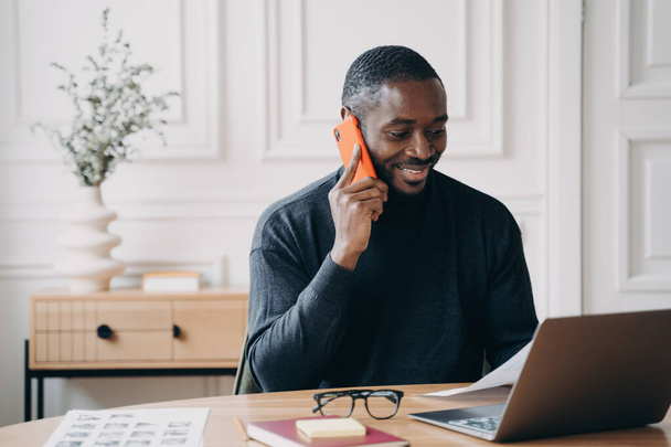 Pozitif Afro-Amerikan erkek müteahhit masada oturmuş bilgisayar ekranına bakıyor, müşteriyle akıllı telefondan konuşuyor, ev-iş meselelerinden çalışan uygulamaları uzaktan tartışıyor. - Fotoğraf, Görsel