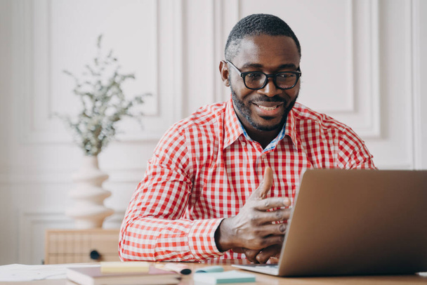 Młody, uśmiechnięty afrykański biznesmen w okularach rozmawiający z partnerem biznesowym przez wideo połączenie na laptopie, omawiający wyniki pracy podczas spotkania online z kolegami - Zdjęcie, obraz