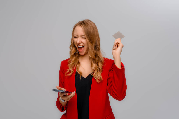 Mulher corporativa feliz segurando smartphone e cartão de crédito, olhando animado sobre venda de loja, pagando on-line, ordem no telefone móvel, fundo cinza - Foto, Imagem