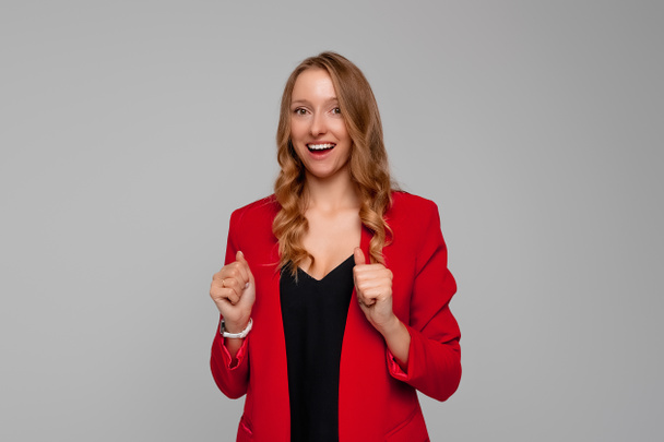 Glad optimistic businesswoman clenches fists for good luck, anticipates positive results, smiles broadly, shows white teeth, dressed in red blazer, expresses happiness - Photo, Image